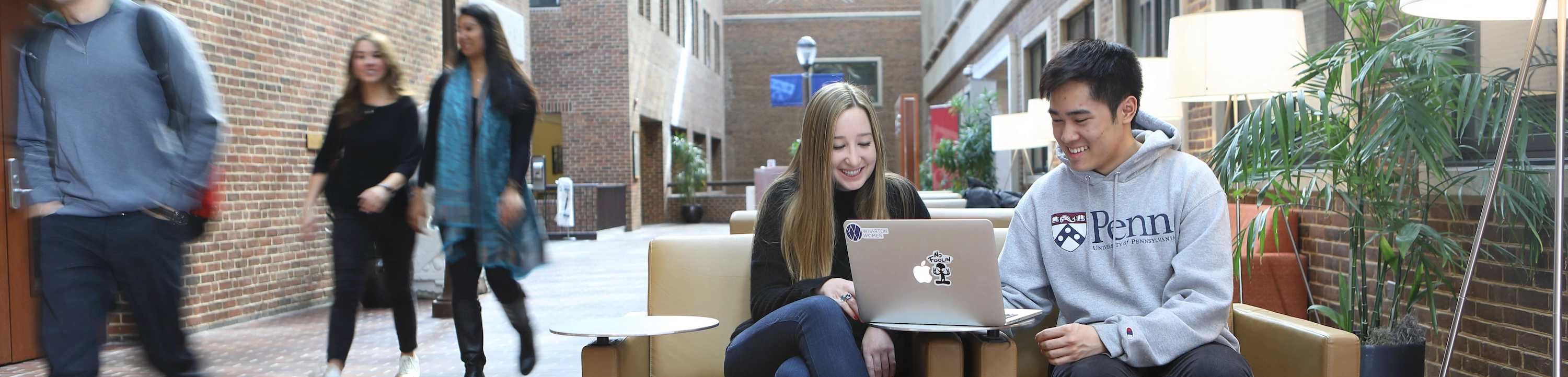 A few students in casual attire at a university indoor common area. Two of them are seated, smiling, and working on a laptop. Others are walking in the background.